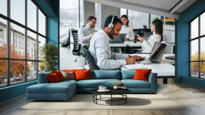 Man is sitting by laptop in headphones. Four people are working in the office together Wall mural