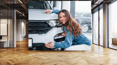 Positive woman in blue shirt embracing her brand new car. In auto salon Wall mural