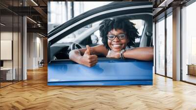 Shows thumb up. Young african american woman sits inside of new modern car Wall mural