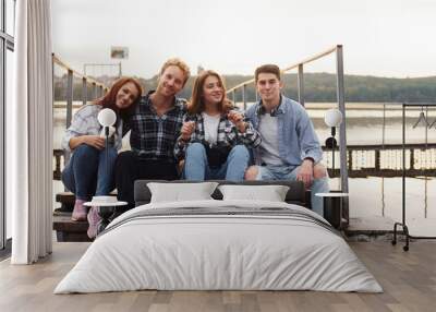 Sitting near lake and embracing each other. Group of young cheerful friends that is outdoors having fun together Wall mural