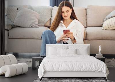 Smartphone in hands. Young woman in white shirt and jeans is at home Wall mural