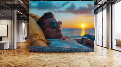 Stylish young man with sunglasses laying on beach couch under blue clear sky on the background of the ocean and sunset Wall mural