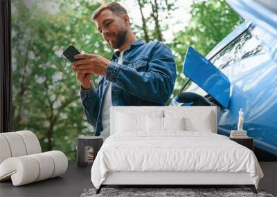 View from below, with smartphone. Handsome man is waiting for his electric car to charge, outdoors Wall mural