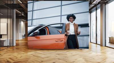 With cup of drink. Young african american woman in glasses stands outdoors near modern car Wall mural