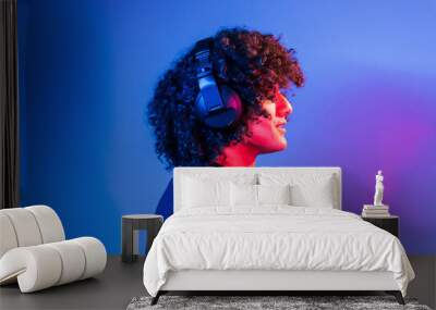 With headphones. Young beautiful man with curly hair is indoors in the studio with neon lighting Wall mural