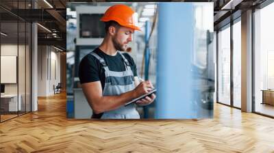 With notepad in hands. Industrial worker indoors in factory. Young technician with orange hard hat Wall mural