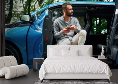 With smartphone in hands. Beautiful young man is sitting in the blue car outdoors Wall mural