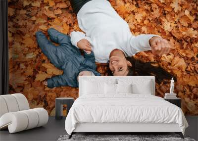 Woman with her little son is laying down on the ground that is covered with autumn fallen leaves Wall mural