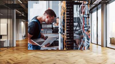 Young man in uniform and with laptop works with internet equipment and wires in server room Wall mural