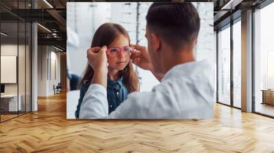 Young pediatrician in white coat helps to get new glasses for little girl Wall mural