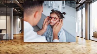 Young pediatrician in white coat helps to get new glasses for little girl Wall mural