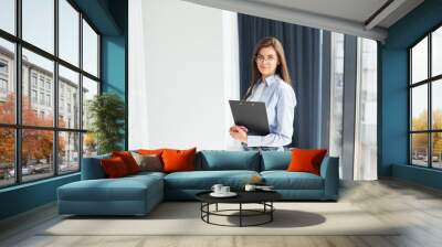 Young woman in glasses, formal clothes and with notepad standing indoors Wall mural