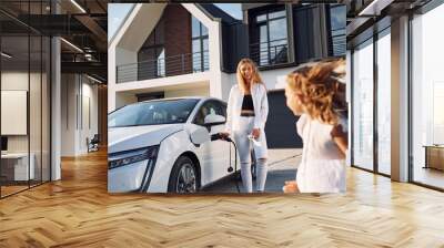 Young woman with her little daughter is with their electric car outdoors Wall mural