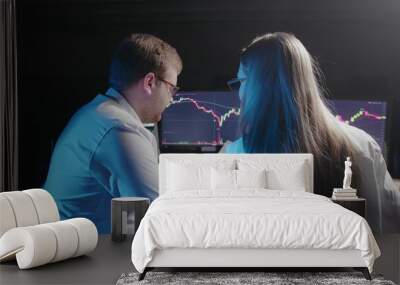 A rear view of a girl and a man sitting at a computer and looking at an online stock market chart showing bitcoin currencies. Two people compare data from paper and computer. In real time Wall mural