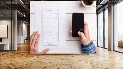 Designer tests the user interface and user experience on a mobile phone. Wireframe beside with mobile phone with a sketched app. The concept of designing software, apps and websites. Wall mural