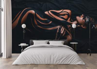 Beautiful girl with painted body and black mask in her face lies on bed. Body art. Wall mural