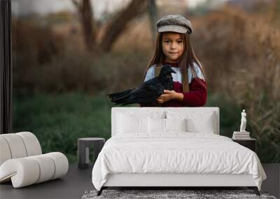 Child girl stands with raven in her hands on background of green grass. Wall mural