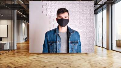 Contagious young male in denim jacket and medical mask looking at camera while standing against white wall Wall mural