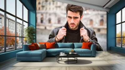 One handsome young man in urban setting in modern city, standing, wearing black leather jacket and jeans, looking at camera Wall mural