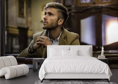 young blond elegant man sitting in church, on wooden bench, thinking and praying, looking around Wall mural