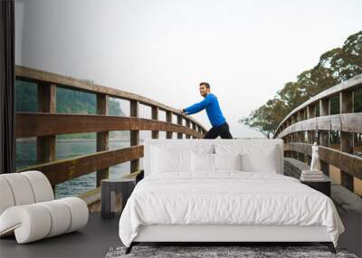 Fit sports man stretching legs before an outdoor running training on a cloudy autumn day with a sport bag on a wooden walkway. Fitness sporty man in Rodiles, Asturias. Wall mural