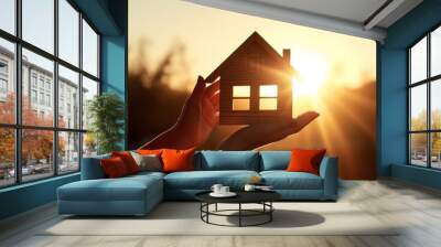 A symbol of your cozy home against the backdrop of the setting sun. Little wooden house in female hand delicately conveying dreams and hopes for secure. Wall mural