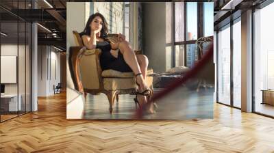 Elegant beautiful woman in a black cocktail dress and high heels sitting in a contemporary armchair in the sunset light Wall mural