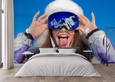 Funny portrait of a young woman in a ski goggles showing tongue on a ski resort. Wall mural