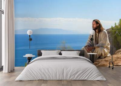 Jesus in robe sitting on stone shore strewn with bushes by sea looking at landscape. Jesus Christ walking near sea. Jesus Christ calmly walking near sea searching for asks for important questions Wall mural