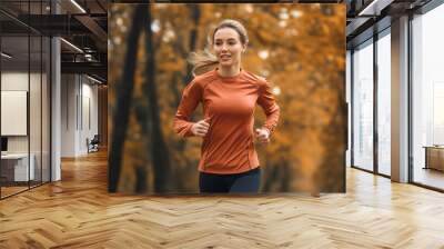 Happy smiling beautiful Caucasian woman running or jogging on trail in autumn park or forest during fall season, female athlete, runner or jogger training or exercising in nature, fitness and sport Wall mural