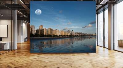 City of Santos, Brazil. Late afternoon on Santos beach with seaside buildings lit by the sun against the blue sky with a full moon. Wall mural