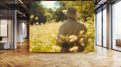 Rearview of an adult mature Caucasian man dressed in a long-sleeve shirt and wearing a straw hat, walking through a sunny field with tall, uncut grass or flowers, enjoying the scenic nature landscape  Wall mural