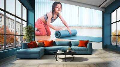 Adorable Asian girl doing yoga exercise, smiling cute child sitting on floor and rolling up yoga mat at indoor studio, sporty kid practicing yoga, working out and exercising for taking care of body. Wall mural