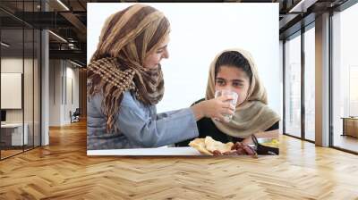 Adorable Pakistani Muslim girl sits at kitchen table with traditional Islamic halal food, kid with hijab drinks water from mother hand on white background, mom feed child daughter, warm love in family Wall mural