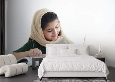 Adorable smiling Pakistani Muslim girl with beautiful eyes wearing hijab, studying online, doing homework by using laptop computer, student kid reading book on white background. Wall mural