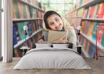 Adorable smiling Pakistani Muslim student girl with beautiful eyes wearing hijab, standing and holding many books with blurred background of bookcase in library or bookstore. Wall mural