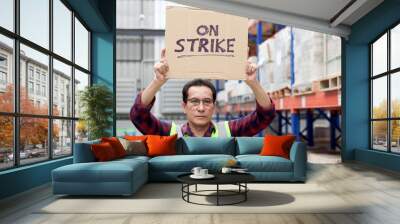 Angry unhappy Asian senior worker wearing safety vest, holding and raising sign on strike banner placard at cargo logistic warehouse. Striking worker protesting at workplace. Wall mural