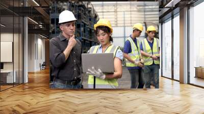 Asian woman worker with safety vest and helmet holds laptop, reports information to her supervisor senior foreman at manufacturing plant factory. Industrial engineer team working together in industry. Wall mural