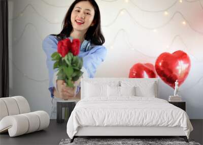 Beautiful happy Asian woman holding red rose bouquet and showing to camera while standing in decorative room. Romantic girl with flower for celebrating love on Valentine Day. Wall mural