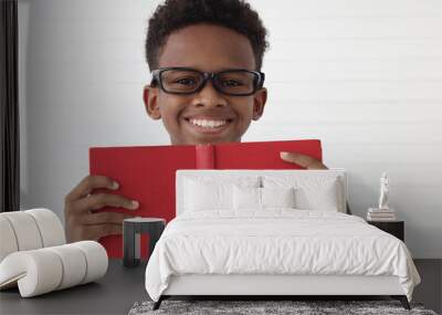 Happy smiling African boy wearing glasses with opening red book on white wall room. Portrait of cute child with book, kid education, reding, learning and studying concept. Wall mural