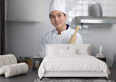 Happy smiling Asian man in white chef uniform with hat, holding wooden rolling kneading stick, standing at kitchen, baker man preparing to bake bread. Portrait of young mam cooking meal food at home. Wall mural