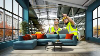 Industrial foreman and worker with helmet and safety vest using laptop computer during discussing together at manufacturing industry factory. Engineer inspecting workplace and checking process. Wall mural