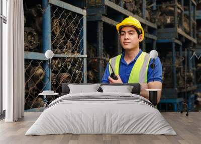 Portrait of industrial engineer worker man wearing helmet with arms crossed, holding radio walkie talkie and standing at manufacturing plant factory with many engine parts as blurred background Wall mural