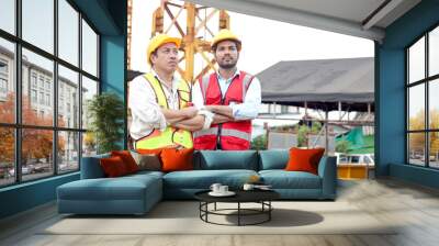 Portrait of two workers with safety vest and helmet standing with arms crossed discussing at construction site. Senior mature elderly engineer and young labor working together at building site. Wall mural