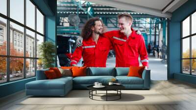 Two handsome mechanic men in red uniform hugging and walking through row of vehicle at garage, auto mechanic technician friend working together to repair customer car automobile at car service shop. Wall mural