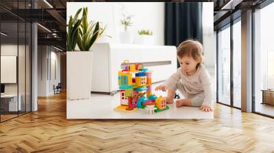 Baby girl playing indoors sitting on floor in playroom at home with constructor. Educational game for baby and toddler. Children build toy house. Creative Wall mural