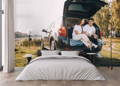 Happy traveler couple sitting in car open trunk and watch sunrise Wall mural