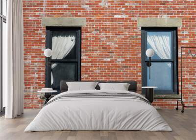 Two windows in a red brick wall. Window ledges are concrete, frames are wooden, painted black. Windows have white curtains, pulled right in one window and left in the other. Wall mural