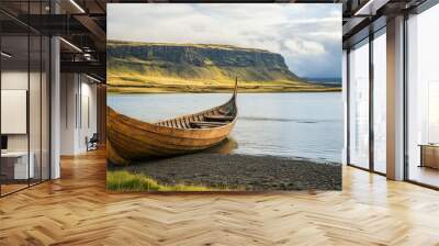 A wooden Viking longship resting on the shore of a tranquil lake, with stunning mountain scenery in the background under a partly cloudy sky. Wall mural