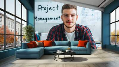 Young Caucasian male in casual attire, confidently smiling while seated at a desk in an office with a 'Project Management' poster in the background. Wall mural